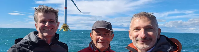 Trois hommes souriants en mer, devant une éolienne flottante