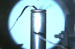 Cellule de batterie cylindrique reliée à des câbles, projetant des particules lors d'un essai