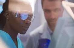 Scientifique en lunettes de protection observant une expérience, un collègue en blouse blanche en arrière-plan