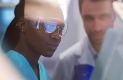 Scientifique en lunettes de protection observant une expérience, un collègue en blouse blanche en arrière-plan