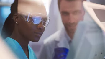 Scientifique en lunettes de protection observant une expérience, un collègue en blouse blanche en arrière-plan