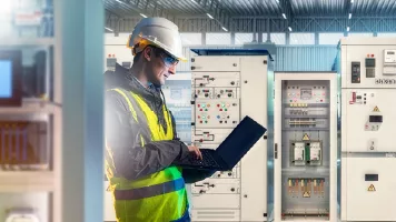 Technicien en casque et gilet de sécurité travaillant sur un ordinateur portable devant des armoires électriques